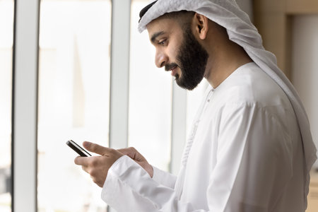 Positive young Arab businessman in white male Muslim clothes using wireless Internet communication for business, typing on smartphone, browsing social media, chatting online. Side viewの写真素材