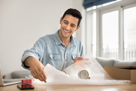 Cheerful handsome Latin customer man wrapping ceramic mugs, packing fragile dish into cardboard box, sitting at workplace table at home, enjoying logistic, transportation routineの写真素材