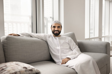 Positive handsome Arabic man in white traditional cloth relaxing at home, sitting on comfortable...