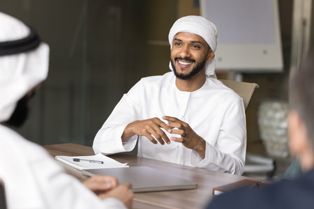 Happy handsome young Arabian business man talking to diverse colleagues on meeting, smiling, laughing, enjoying corporate communication, teamwork, brainstorming on creative ideas. Candid portraitの写真素材