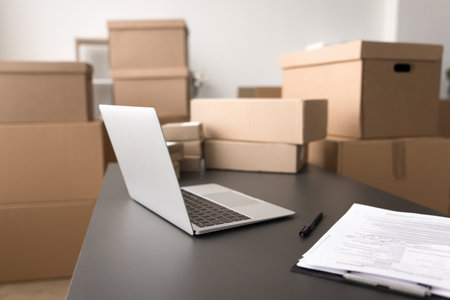 Close up workspace with open laptop, stack of papers on table, multiple cardboard boxes of various...