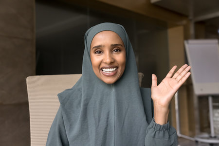 Happy beautiful young Arabian businesswoman in grey hijab talking on conference call from office, waving hand hello at camera, speaking with toothy smile. Video chat screen head shot portraitの写真素材