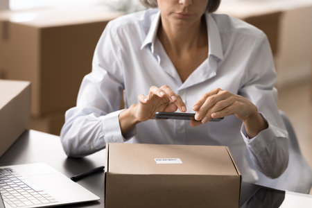 Close up woman sits at desk, hold phone scanning barcode, track packages with mobile bar code tech, optimize delivery process, updating shipping status, confirm package details, enters inventory dataの写真素材