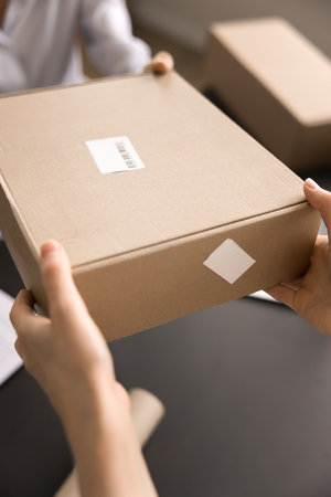 Close up view unknown female hands giving, receiving cardboard package. Client takes box in postal services. Workflow of post office or warehouse worker. Parcel shipping management, orders fulfilmentの写真素材