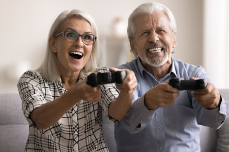 Amazed happy aged married couple having fun at home, holding video game controller, fighting in virtual battle, laughing, playing videogames with excitement and passion. Indoor activity, entertainmentの写真素材