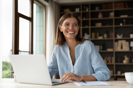 Cheerful young female worker sitting at workplace with laptop in office, looking away, smiling, laughing. Millennial employee, business woman in casual distracting from work, talking to colleaguesの写真素材