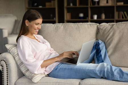 Happy young woman resting on comfortable couch, using laptop computer, typing, looking at screen, smiling, laughing. Cheerful girl chatting online on social media, reading messages, browsing internetの写真素材