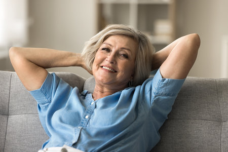 Relaxed mature woman looking at camera lying indoor leaned on sofa with hands behind head, spend carefree retired life, day off or weekend leisure alone at home. Pastime, pleasant rest in living roomの写真素材