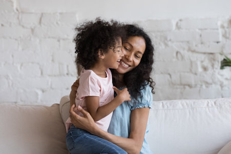 Young caring African mother sit on sofa hugging cheerful laughing little 5s daughter, loving family enjoy time together, embracing feeling unconditional love. Happy motherhood, bond, custody conceptの写真素材