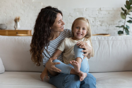 Little 5s cute blond daughter sits on loving Hispanic mothers laps smile looking at camera, enjoy time, tender moments together seated on sofa at home. Happy family, adoption, bonding, cherish conceptの写真素材