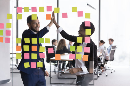 Excited diverse business partners celebrating successful teamwork, completing project, goal achieving, giving high five at class board with paper sticky notes, smiling, laughing. Through glass viewの写真素材