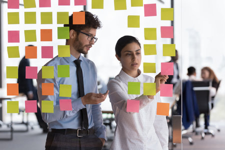 Diverse millennial employees, coworkers discussing project schedule, writing memo on sticky notes....