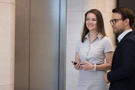 Confident coworkers standing in office hall, waiting for elevator, chatting, smiling, looking away....
