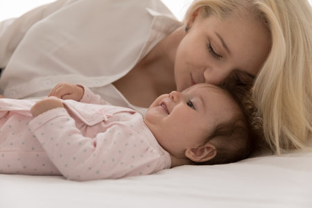 Young loving mother kisses on forehead her lovely babygirl, lying together on bed, close up cropped shot. Woman feeling pure love to newborn baby, express caress. Happy motherhood, family bond conceptの写真素材