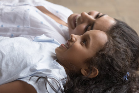 Cute little Indian girl and loving smiling mother lying together on floor indoors, close up. Young woman spend leisure with preschooler daughter, enjoy carefree time and happy motherhood. Love, bondの写真素材