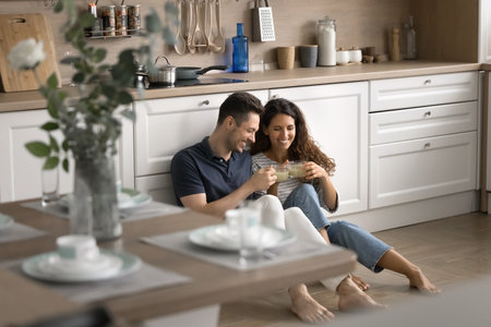 Cheerful positive young romantic couple sitting on heating floor in modern kitchen, toasting mugs with drink, having fun, laughing, enjoying leisure, break at new homeの写真素材