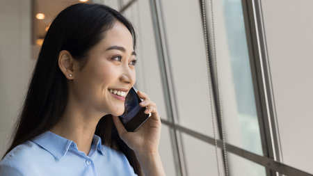 Cheerful beautiful young Asian employee woman speaking on mobile phone at large office window, looking away with toothy smile, enjoying telephone call, conversation, communication. Banner shotの写真素材