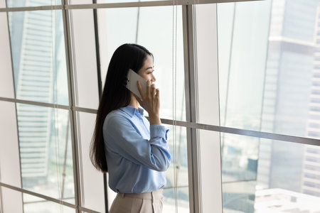 Thoughtful young Asian business woman talking on cellphone in office space, standing at large window, looking away at city view, thinking on conversation, commercial communicationの写真素材