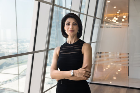 Successful confident young brunette business leader woman looking at camera with hands folded, standing in modern urban office workspace at large window, posing for professional portraitの写真素材
