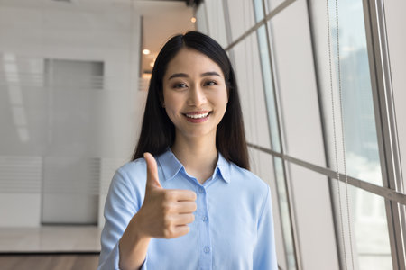 Happy young Asian business professional girl making thumb up hand, showing like gesture at camera, smiling, laughing, posing for portrait, expressing approval, giving positive feedbackの写真素材