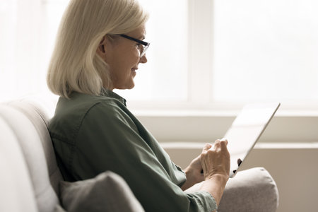 Mature woman wear glasses using tablet seated on sofa at home, do online shopping, buying e-services, ordering goods and gifts, accessing to services without leaving home, enjoy convenience and safetyの写真素材