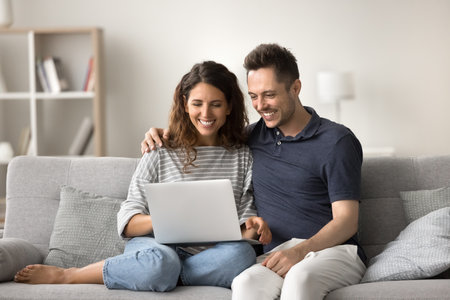 Cheerful attractive romantic couple using computer at home, sitting close on sofa, using modern Internet technology, online app, media service for communication, having fun, laughingの写真素材
