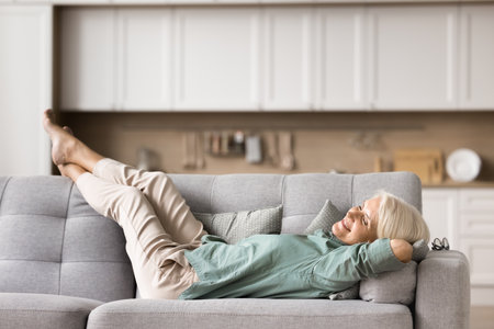 Happy mature woman lying comfortable on cozy sofa at home, put hands behind head enjoy pleasant carefree relaxation alone in studio flat, take break, relish coziness at home. Daytime nap, well-beingの写真素材