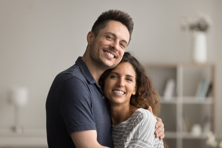 Happy positive young married couple hugging with love, care, enjoying romantic affection, close relationship, standing at home, looking at camera, smiling, laughing, posing for portraitの写真素材