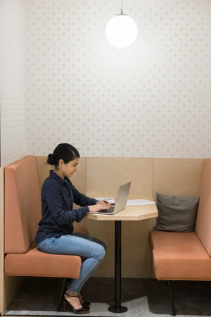 Serious young Indian freelancer girl working at laptop in office booth, typing at table....
