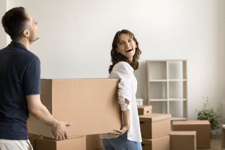 Pleasant chores. Laughing newlyweds husband wife carry carton packages with stuff furniture decor to...