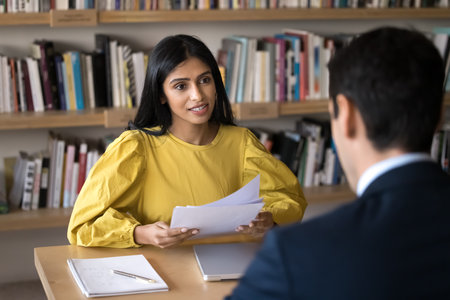 Young Indian recruit manager woman talking to job candidate at interview, holding paper resume. Diverse student and teacher meeting in library, discussing research studyの写真素材