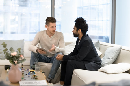 Two multiethnic business colleagues discussing work tasks on office couch, sharing tablet computer, talking. Caucasian businessman showing online startup project to Arab investor, partnerの写真素材