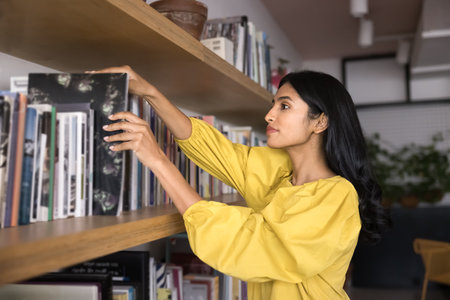 Focused young Adult Indian student woman taking textbook from bookshelf for studying, looking for information in public library, getting knowledge. Customer choosing book in bookstoreの写真素材