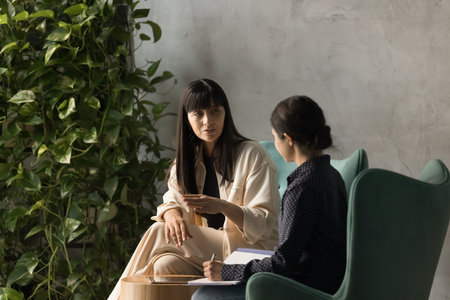Serious middle aged businesswoman, mentor teaching young female intern, sitting in office armchair, speaking for colleague listening and making notes. Indian professional interviewing expertの写真素材