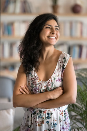 Cheerful young Indian student woman in summer dress posing in library, standing with hands folded,...