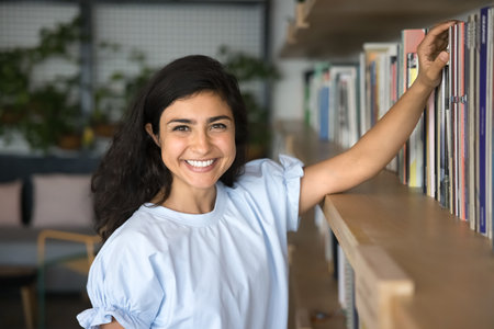 Cheerful beautiful young Indian literature fan woman choosing book on bookshelf, looking at camera...