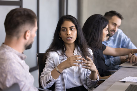 Serious beautiful young Indian professional woman talking to colleague at workplace. Multiethnic business colleagues, project managers talking at work table, discussing project tasks, teamworkの写真素材