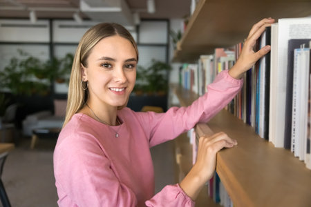Positive young female student picking textbook from shelf, visiting college library, looking at...