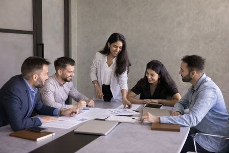 Multiethnic business team of positive young colleagues analyzing marketing data, talking at meeting table, reviewing reports with financial statistic diagrams, working on project strategy togetherの写真素材