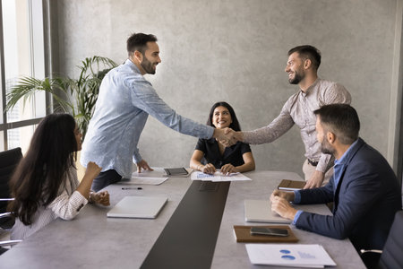 Two positive successful business partners shaking hands over large table. Team of stake holders, project managers, investors ending negotiation meeting with handshake, smiling, laughingの写真素材