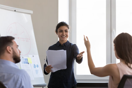 Happy young Indian business coach woman getting question from audience on workshop meeting, pointing at asking employee with raise hand, smiling. Corporate training, presentation conceptの写真素材