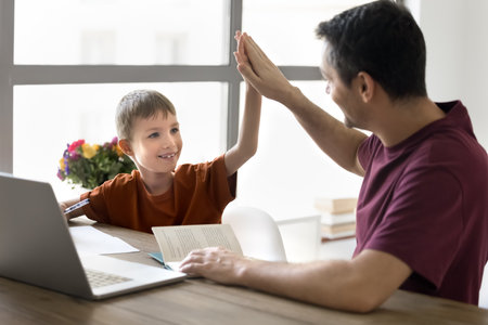 Happy little schoolkid son and proud dad giving high five at home table with desk and copybook,...