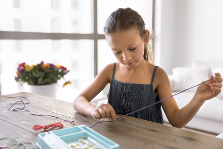 Focused calm silent cute little girl making creative handmade decoration from thread and beads,...