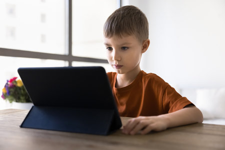 Focused early schoolchild aged boy studying online at tablet computer, using educational learning...