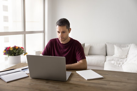 Focused middle aged Hispanic freelance business man in casual t-shirt working on online project at...