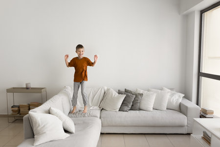 Happy active boy kid jumping on large soft couch in cozy home interior with white wall in background, smiling, laughing, having fun, spending healthy energy. Full length wide shotの写真素材