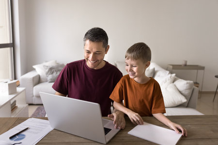 Cheerful Hispanic freelance daddy and positive cute son kid using laptop at home workplace table...