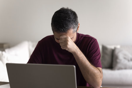 Upset unhappy freelance employee man touching head at laptop, hiding face, working from home, getting failure project result, financial problems, money loss, difficult job taskの写真素材