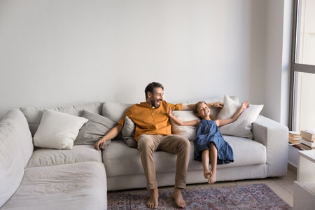 Joyful excited Latin dad and playful little daughter kid resting on spacious comfortable white home couch with open hands, laughing, having fun, enjoying carefree family leisureの写真素材