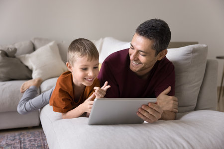 Cheerful dad and son kid using Internet technology at home, watching funny online video content, browsing social media on tablet, discussing application, service, communication, smiling, laughingの写真素材